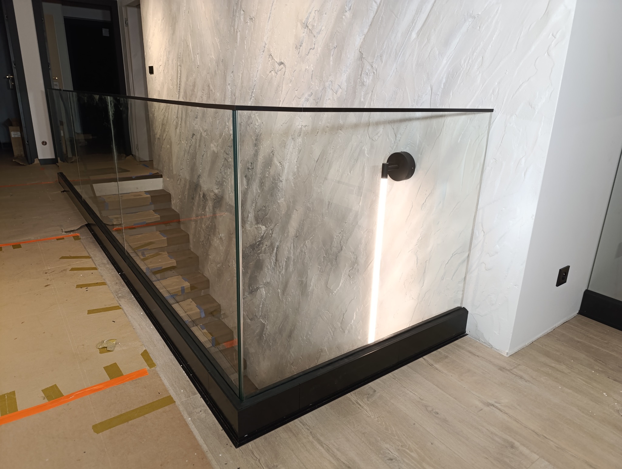 Szklana balustrada z czarną ramą na schodach wewnętrznych, widoczne zabezpieczenia na stopniach, oświetlenie ścienne LED.