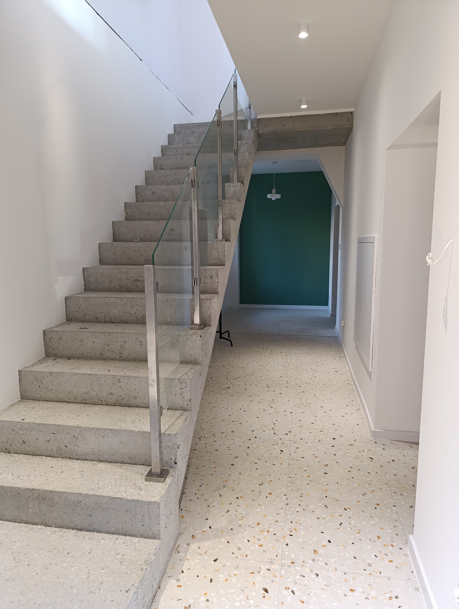 Betonowe schody z szklaną balustradą w nowoczesnym wnętrzu, widok z dołu na górę, z zieloną ścianą w tle.