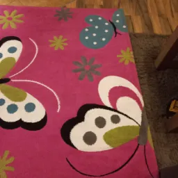 Różowy dywan z motywem motyli i kwiatów leżący na drewnianej podłodze, częściowo zakryty przez szary dywan.