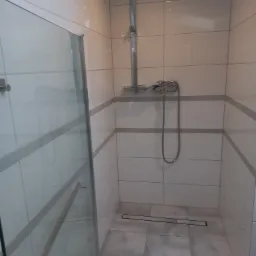 Kabina prysznicowa z szklanymi drzwiami i chromowaną armaturą, wykończona białymi płytkami z szarymi pasami i szarą podłogą z odpływem liniowym.