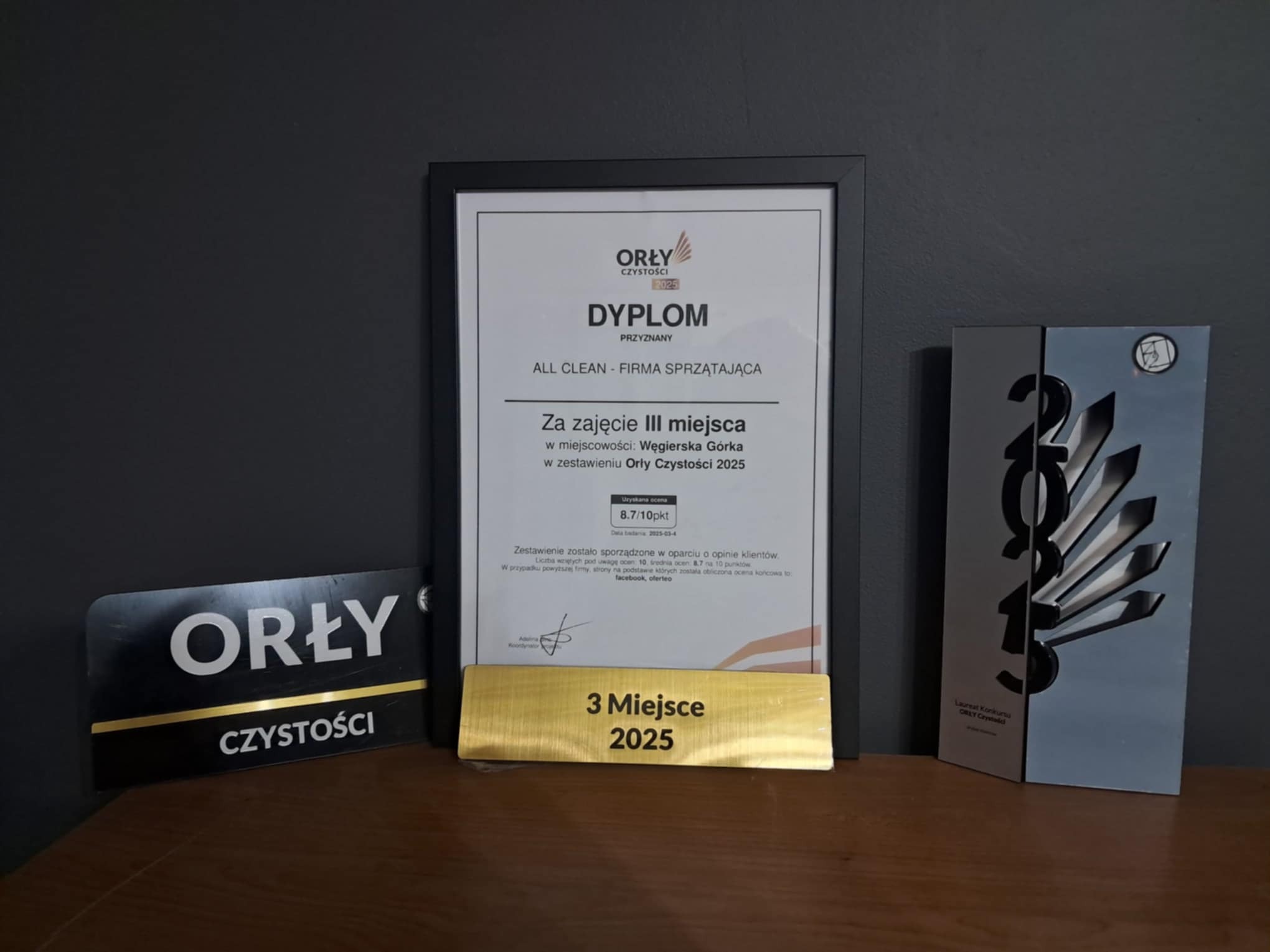 Dyplom 'Orły Czystości 2025' przyznany firmie sprzątającej ALL CLEAN za zajęcie III miejsca, obok statuetka i tabliczka z nazwą konkursu, ustawione na drewnianym blacie na tle szarej ściany.