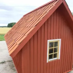 Czerwony, drewniany domek z oknem i dachówką ceramiczną, stojący na betonowej płycie, z widokiem na pole i las w tle.