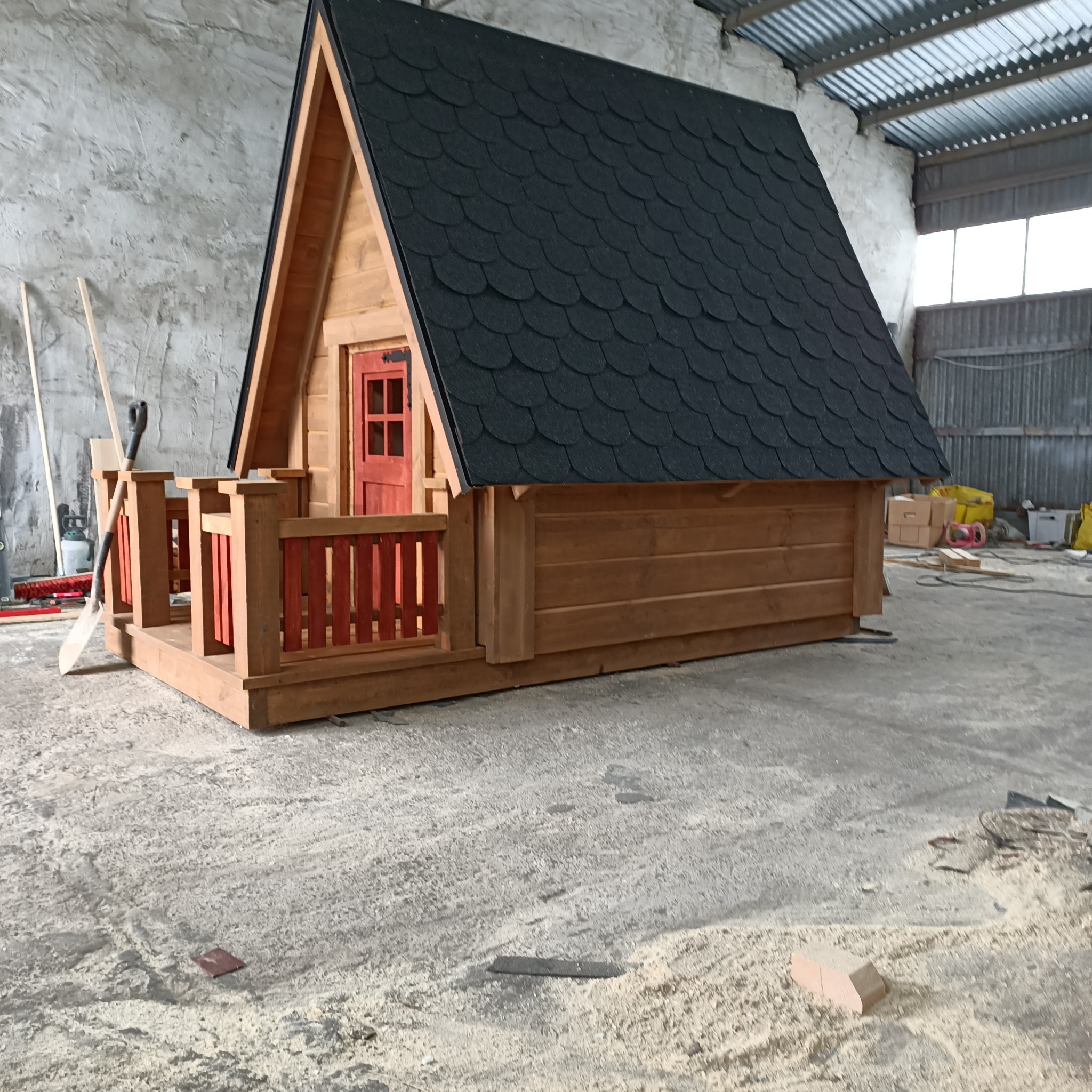 Drewniany domek z czarnym dachem z gontu bitumicznego i czerwoną stolarką okienną i drzwiową, stojący w hali produkcyjnej, z widocznymi narzędziami i odpadami po obróbce drewna.