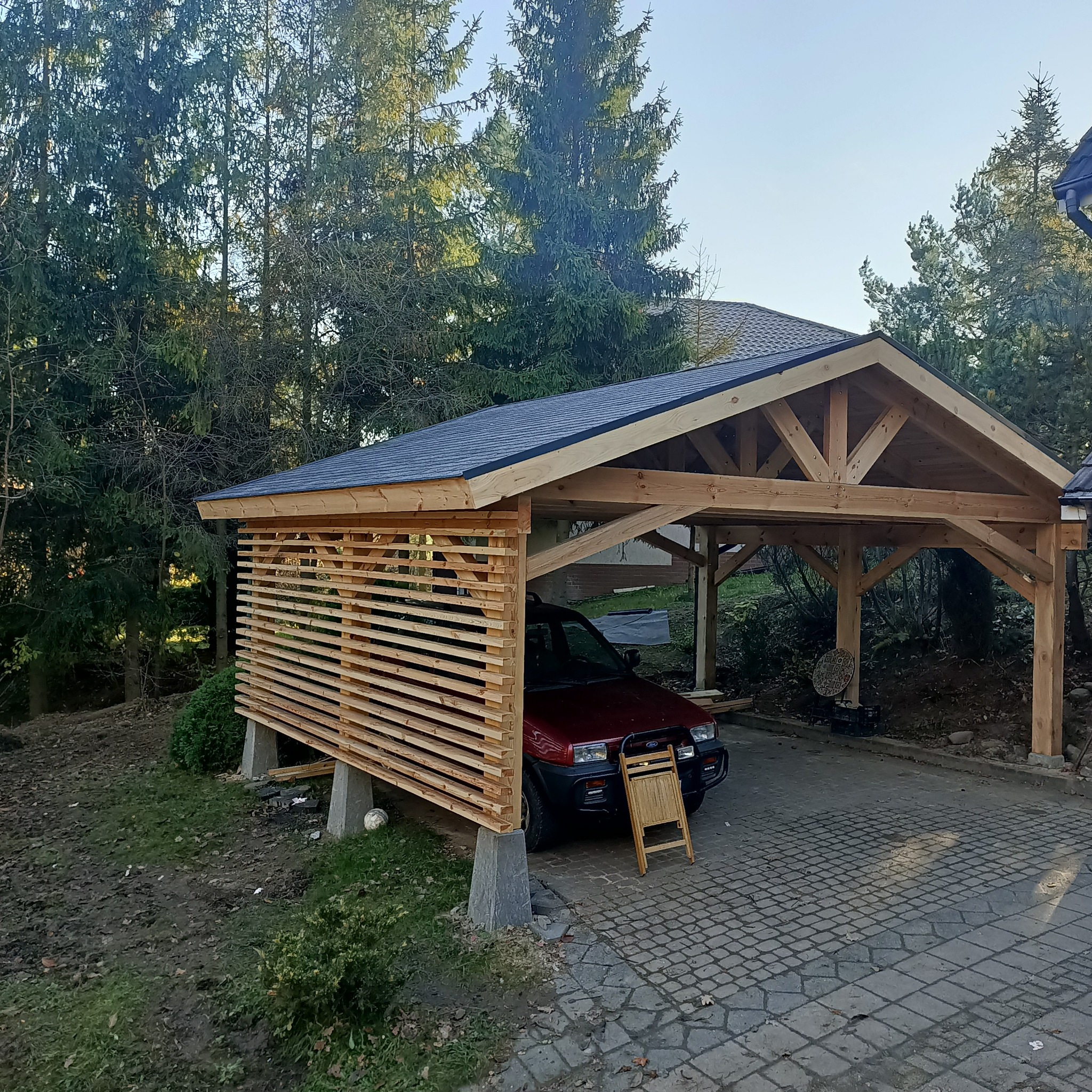 Drewniana wiata garażowa z ażurową ścianą boczną, częściowo zadaszona, z zaparkowanym czerwonym samochodem osobowym i składanym krzesłem z przodu, na tle drzew iglastych.