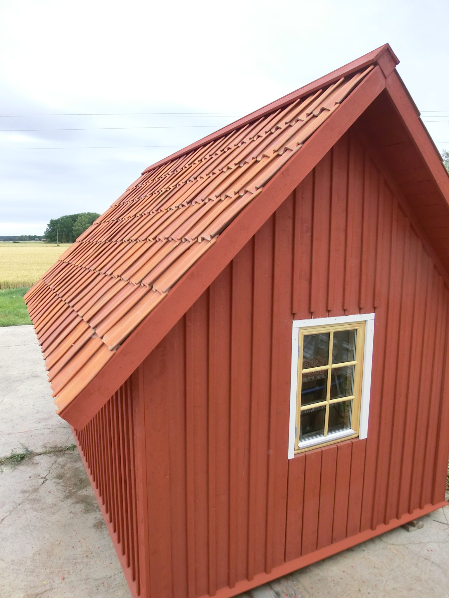 Czerwony, drewniany domek z oknem i dachówką ceramiczną, stojący na betonowej płycie, z widokiem na pole i las w tle.
