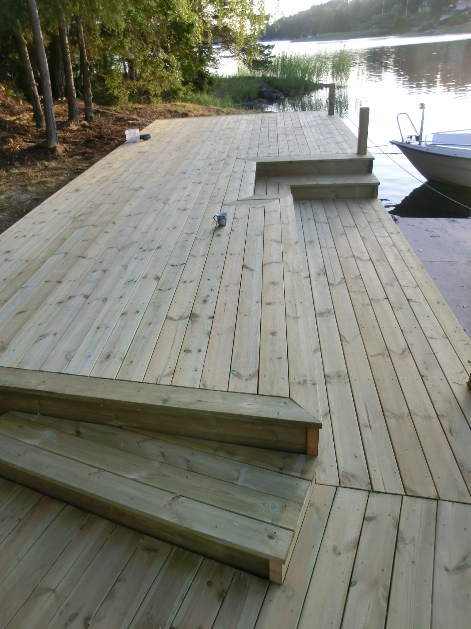 Nowy drewniany pomost z krótkimi schodami prowadzącymi do jeziora, obok zacumowana łódka, w tle drzewa i zachodzące słońce.