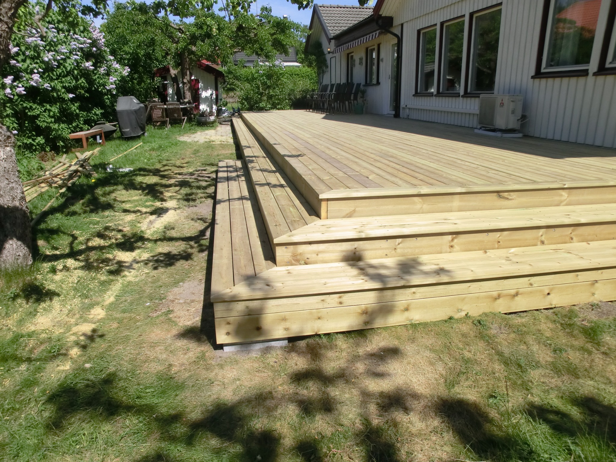 Nowo wybudowany drewniany taras z trzema stopniami, wykonany z jasnego drewna, przylegający do białego domu z ciemnymi ramami okiennymi, widoczny trawnik i cień drzewa.