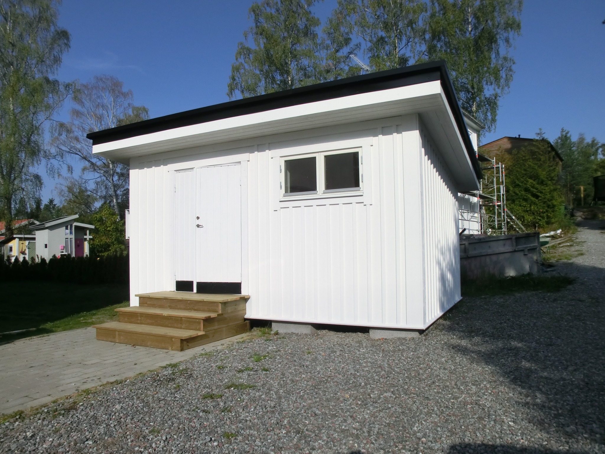 Biały, drewniany budynek gospodarczy z czarnym dachem i drewnianymi schodami wejściowymi, otoczony żwirem i zielenią, na tle błękitnego nieba i drzew.