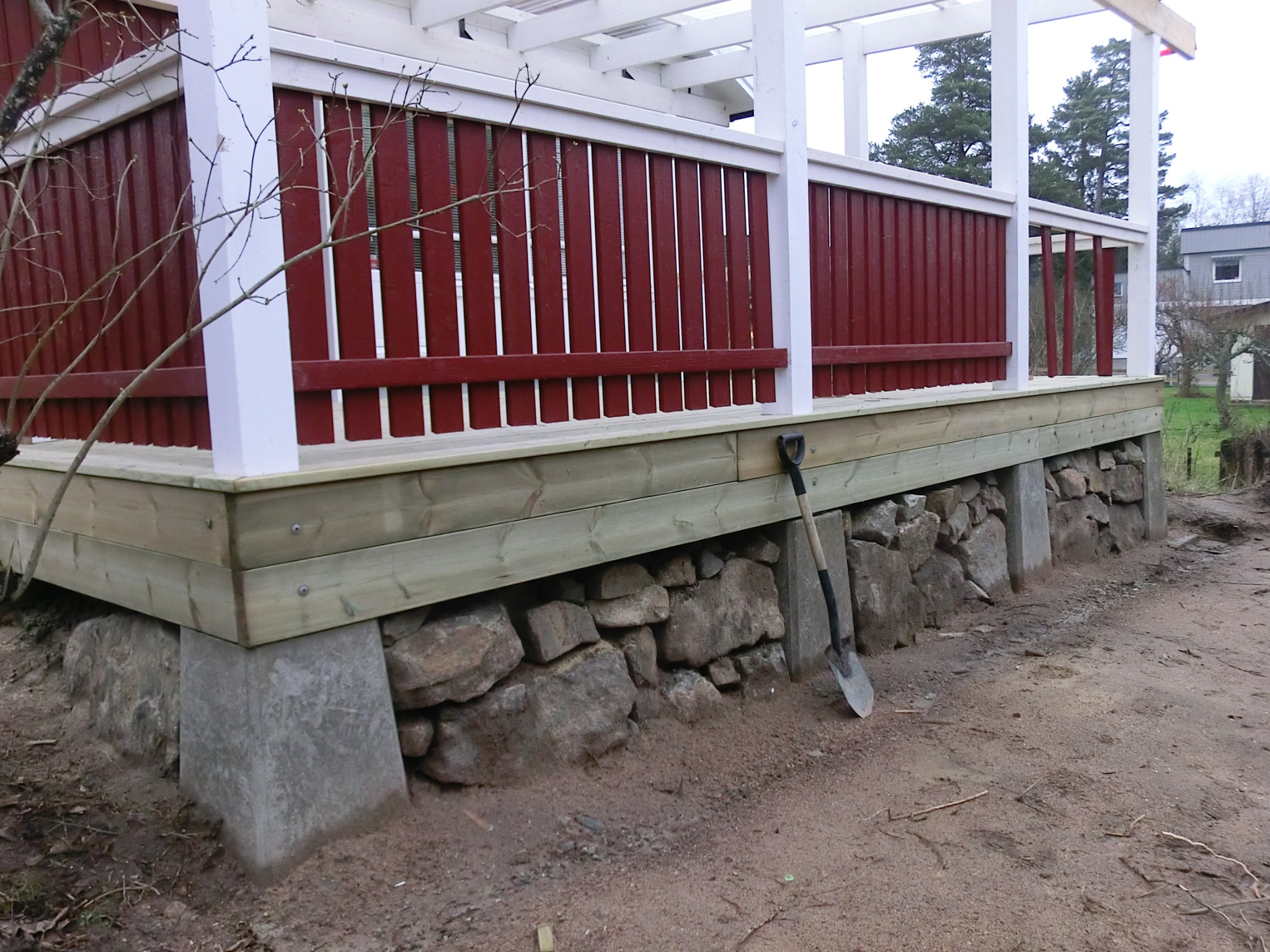 Nowo wybudowany taras z czerwonymi barierkami i białymi słupkami, podparty na betonowych fundamentach i kamiennej podmurówce, z łopatą opartą o bok.