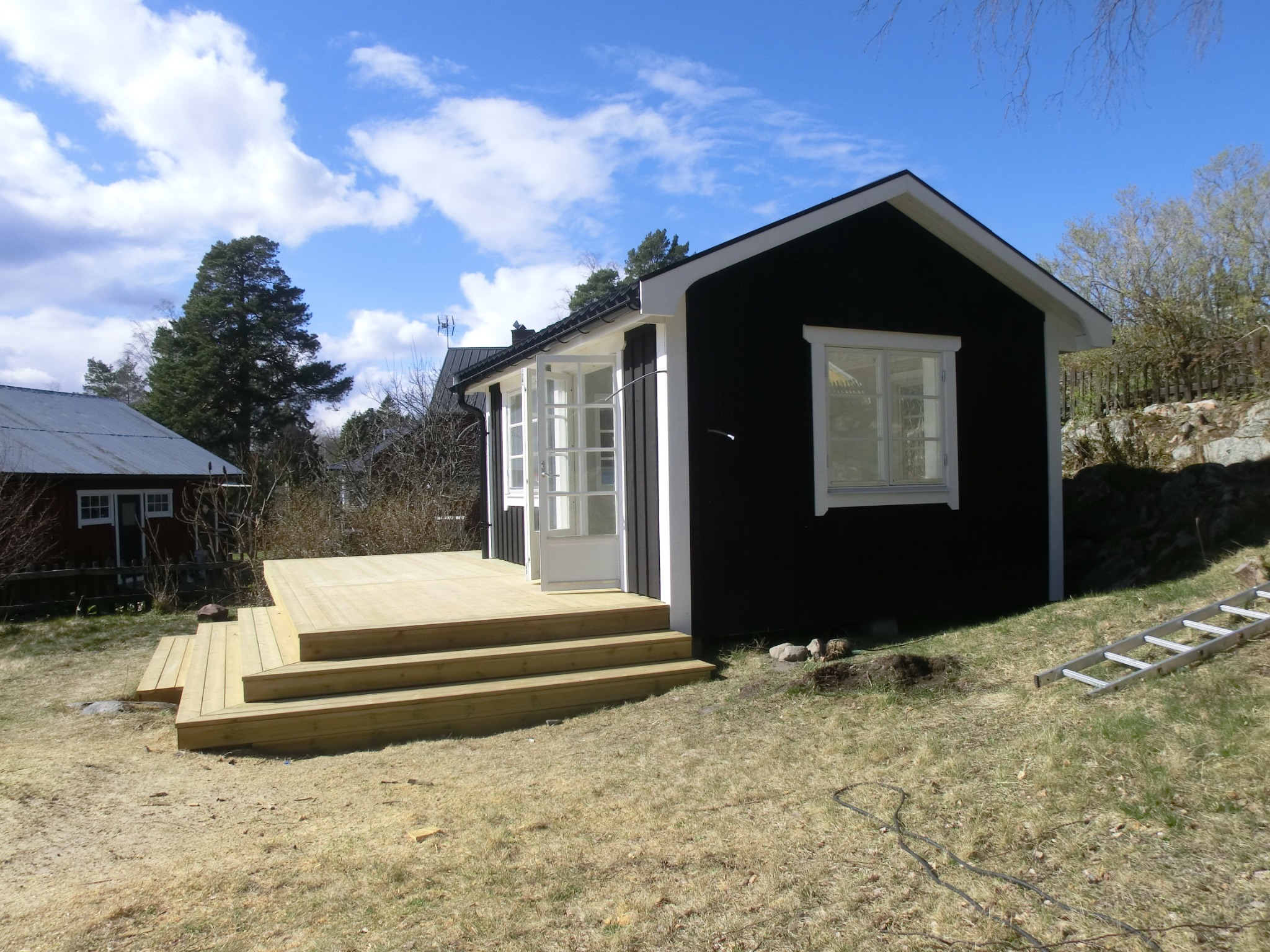 Czarny, parterowy domek z nowo wybudowanym drewnianym tarasem z trzema stopniami, białe ramy okienne i drzwi, na trawniku leży drabina.