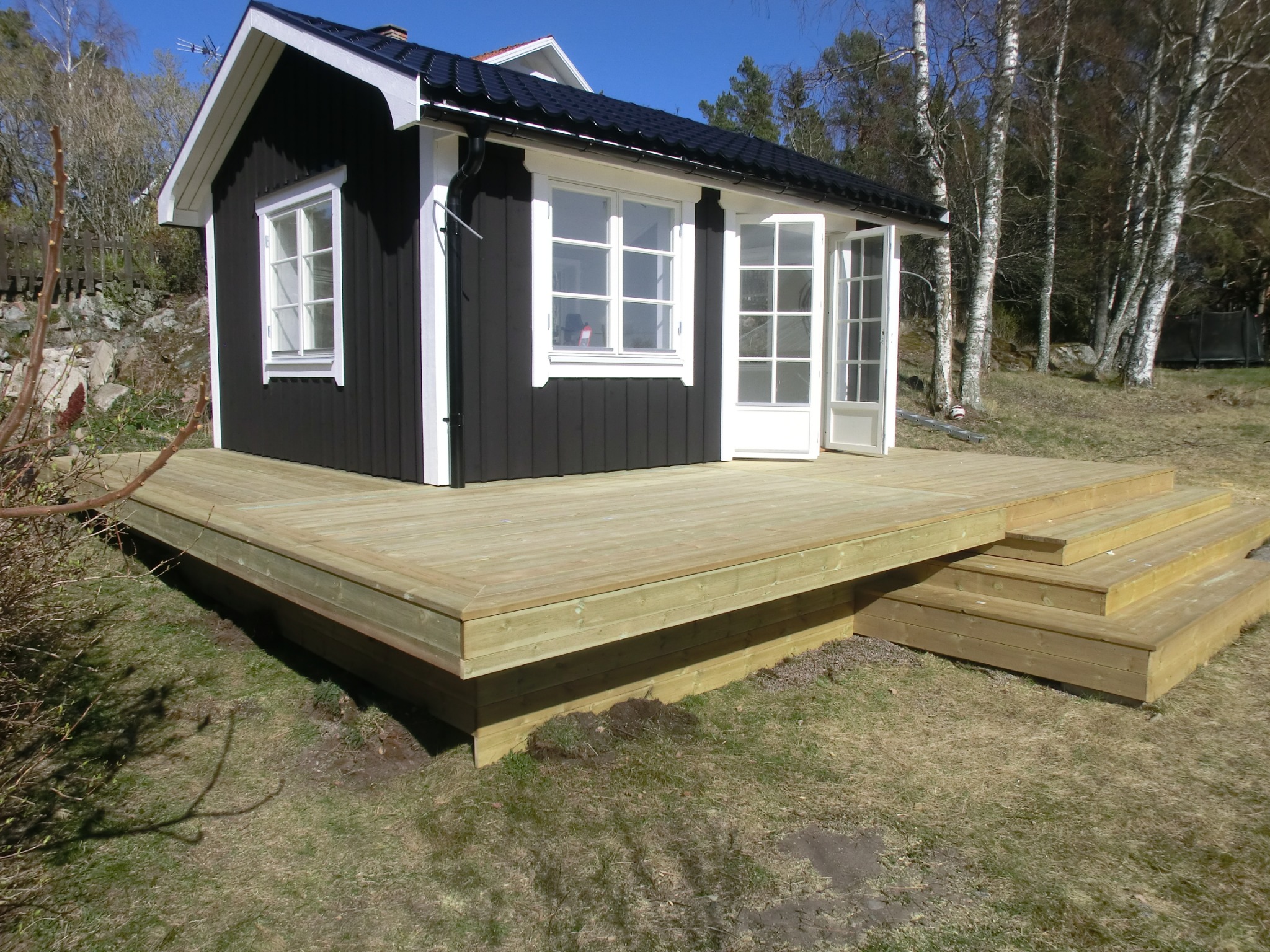 Nowo wybudowany drewniany taras przylegający do ciemnego domku z białymi oknami i drzwiami, schody tarasowe prowadzą na trawnik.