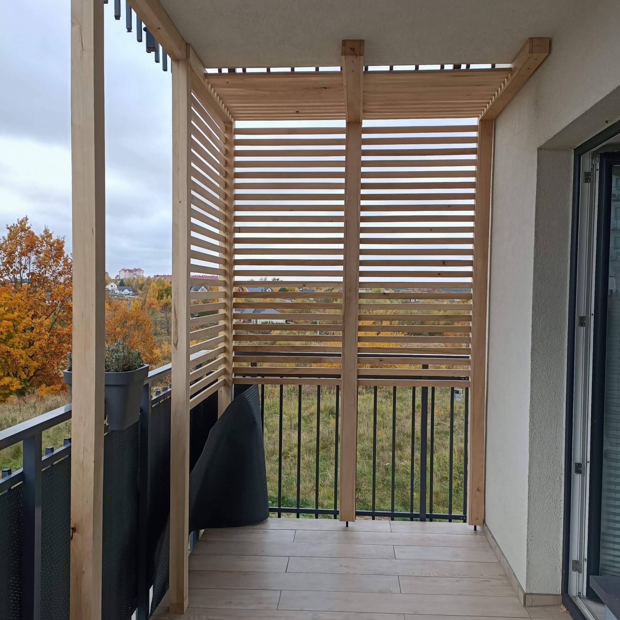 Drewniana konstrukcja z poziomych listew na balkonie, widok na jesienny krajobraz z drzewami w odcieniach pomarańczy i czerwieni, szara donica z rośliną na balustradzie.