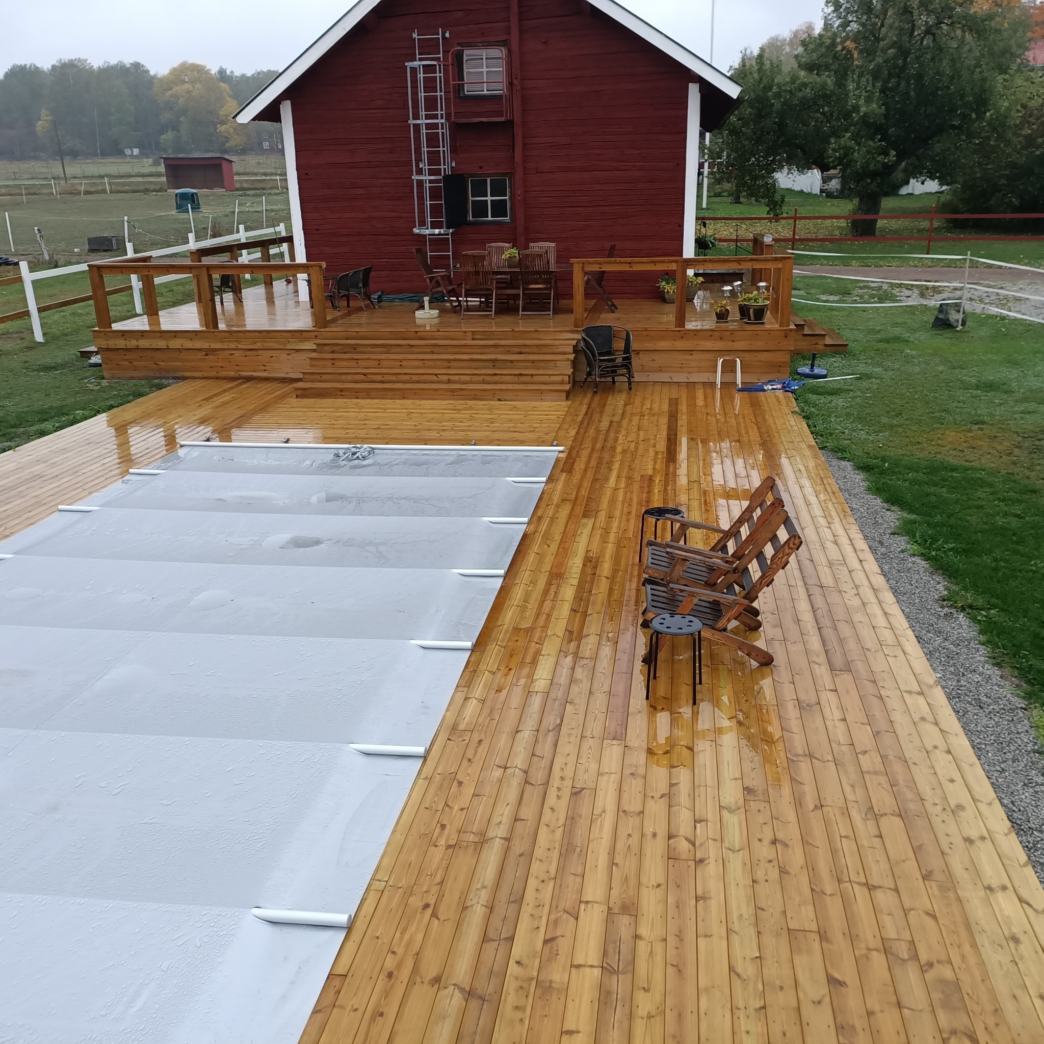 Drewniany taras z widokiem na basen z szarą pokrywą, obok czerwony budynek gospodarczy z drabiną, w tle zielony trawnik i drzewa.
