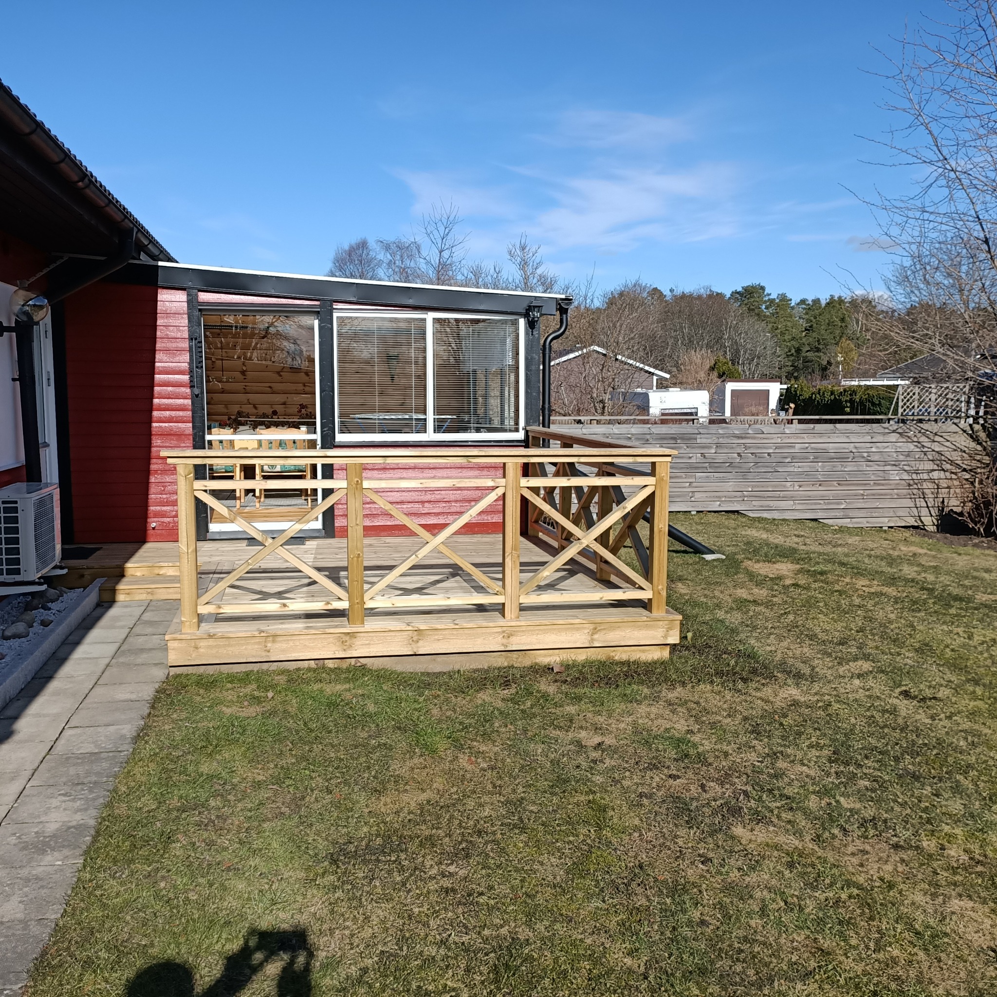 Nowo wybudowany drewniany taras z balustradą w kształcie krzyży, przylegający do czerwonego domu z widocznym klimatyzatorem na zewnątrz, w tle trawnik i drewniany płot.