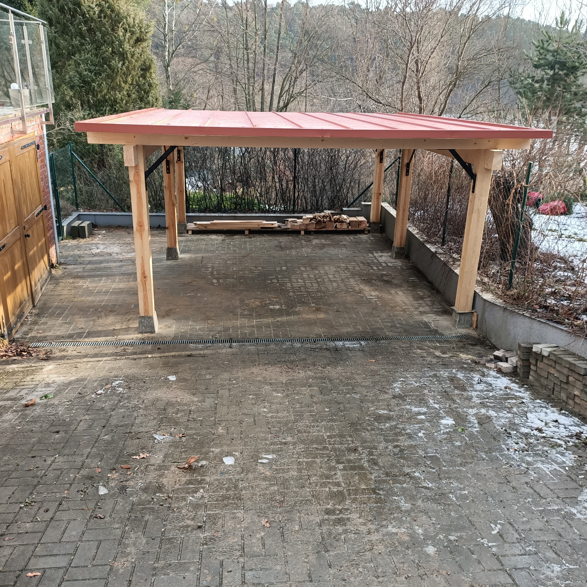Drewniana wiata z czerwonym dachem na posesji, widoczne drewniane elementy konstrukcyjne i przygotowane deski, kostka brukowa na podłożu, w tle drzewa bez liści.