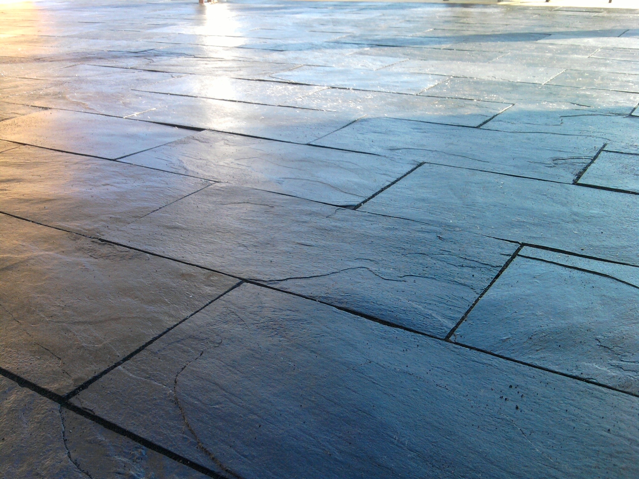 Nowa, ciemnoszara posadzka z płytek imitujących kamień, ułożonych na zewnątrz, z widocznymi fugami i odbijającym się światłem.