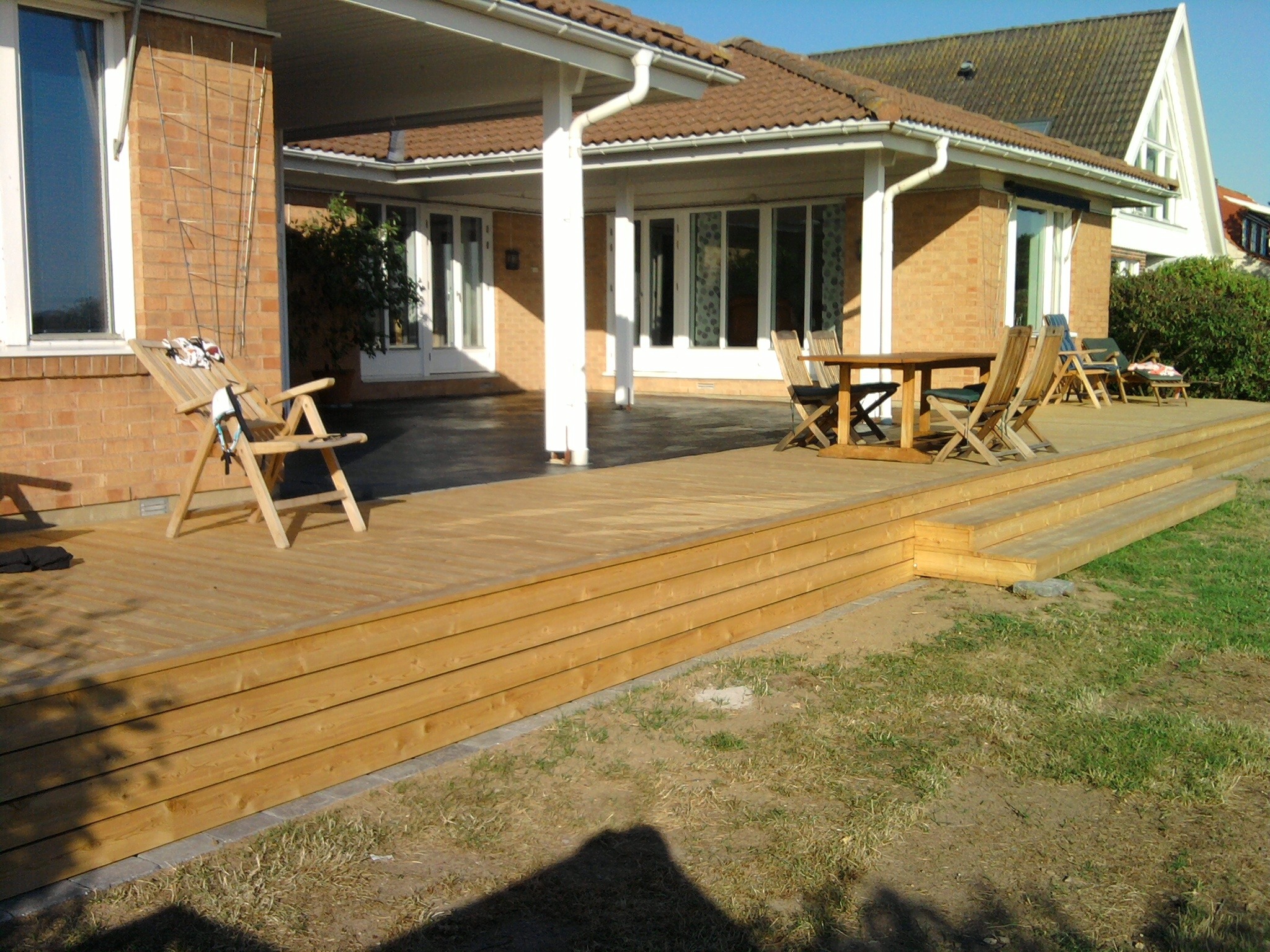 Nowy drewniany taras przylegający do domu, z widocznymi schodami, meblami ogrodowymi i zadaszeniem wspartym na białych kolumnach.