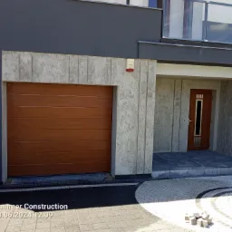 Nowoczesny dom z drewnianą bramą garażową segmentową, betonową elewacją i kostką brukową ułożoną we wzór koła.