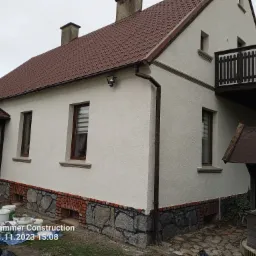 Elewacja domu jednorodzinnego po remoncie w Trzcińsko-Zdrój, z ociepleniem i tynkiem strukturalnym, widoczne fragmenty kamiennej podmurówki i brązowej dachówki, oraz puszki z farbą na pierwszym...