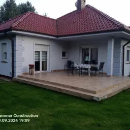 Dom z czerwonym dachem i jasną elewacją, widok z ogrodu. Taras z drewnopodobnych płytek, krzesła i stół na zewnątrz. Uwagę przykuwają detale wykończenia elewacji.