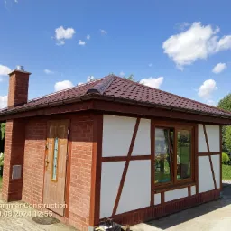 Wykończony budynek w stylu muru pruskiego z brązowym dachem, kominem i stolarką okienną na tle błękitnego nieba z chmurami, otoczony zielenią.