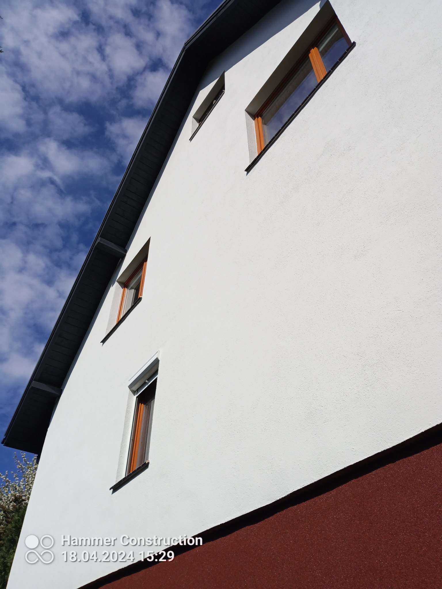 Świeżo otynkowana, biała elewacja domu z brązowym cokołem i drewnianymi ramami okiennymi, uchwycona pod kątem z widocznym fragmentem błękitnego nieba z chmurami.