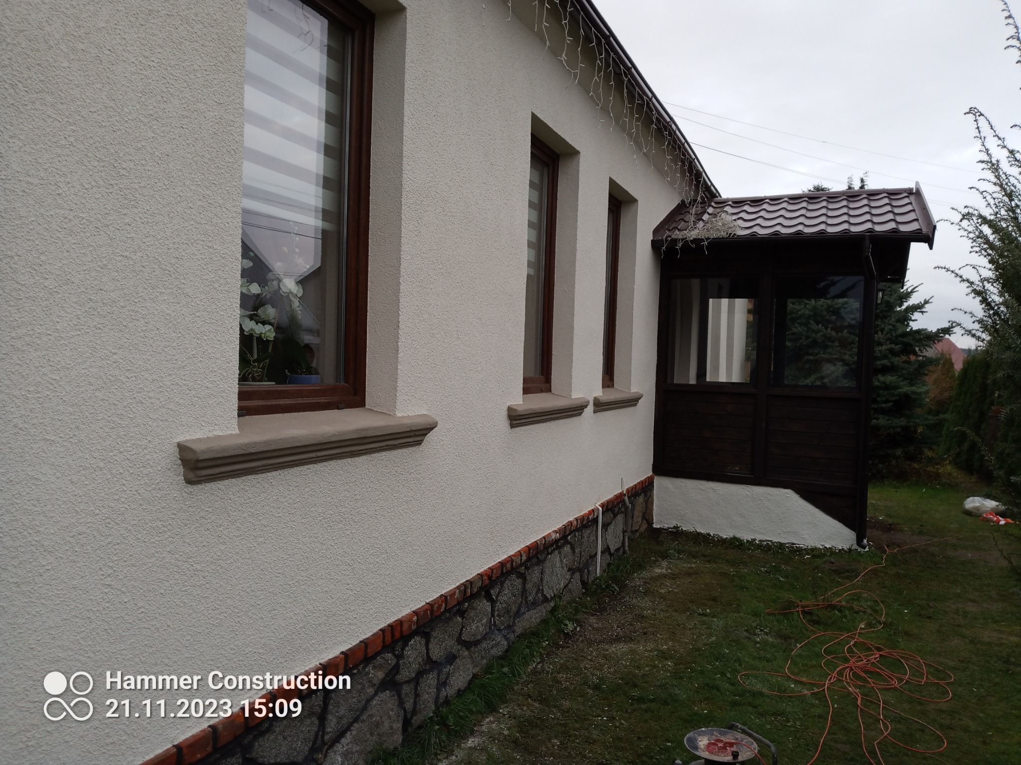 Boczny widok domu po renowacji elewacji z dekoracyjnym tynkiem, widoczne okna z brązowymi ramami, kamienny cokół i dobudowany drewniany ganek z brązowym dachem.