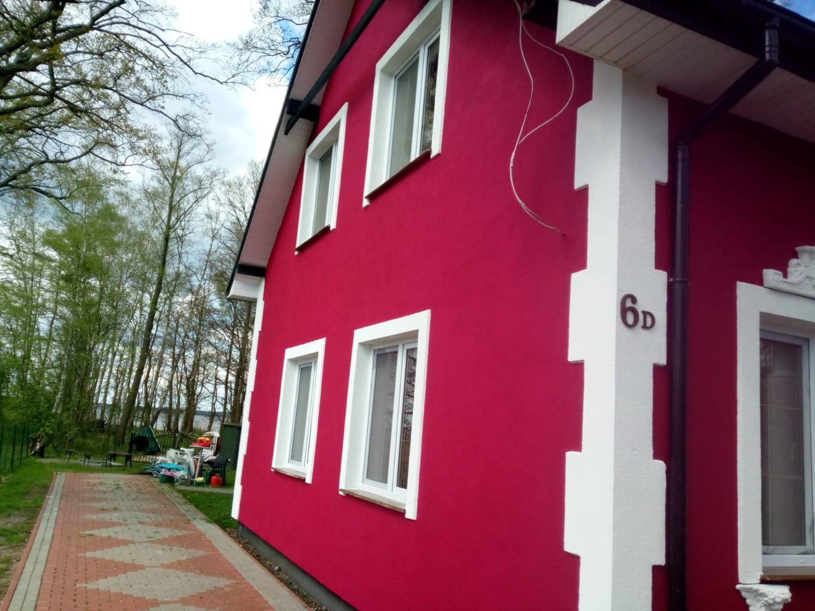 Czerwona elewacja domu jednorodzinnego z białymi obramowaniami okien i narożników, widoczny numer domu '6d', na tle drzew i częściowo widocznego jeziora.