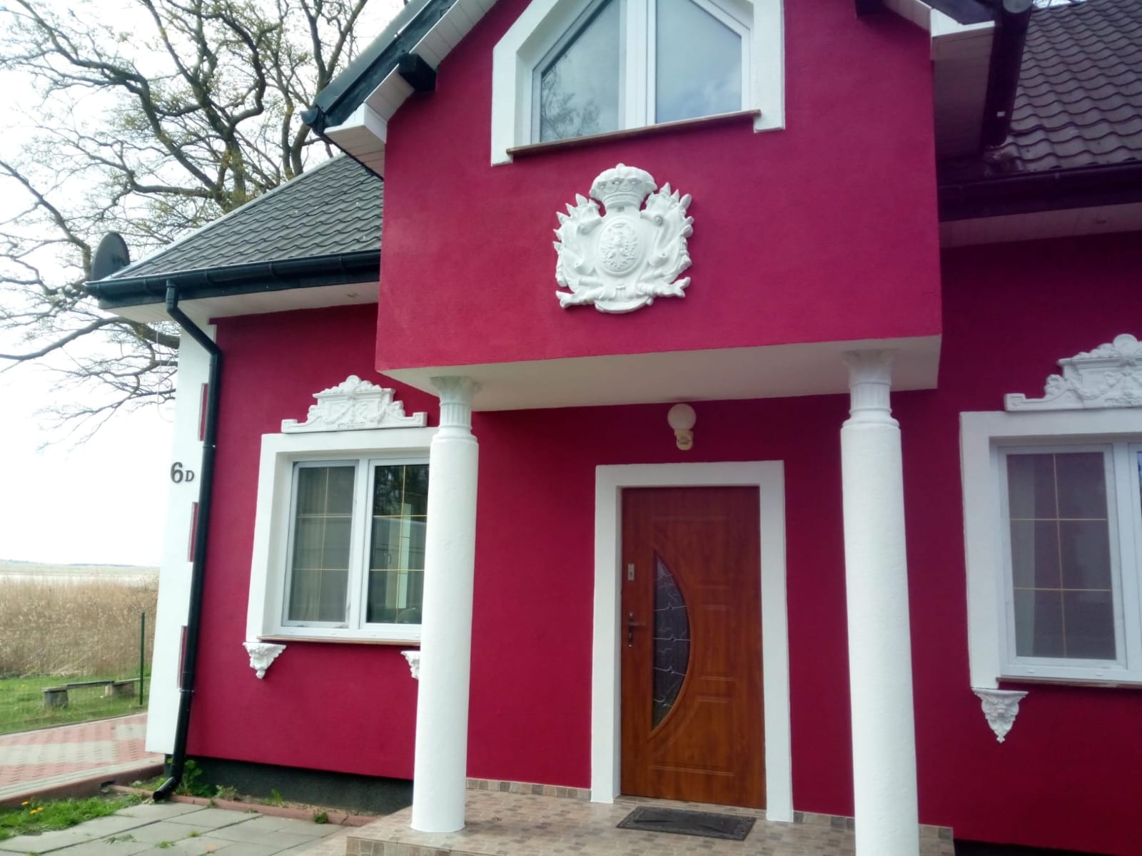 Dom z intensywnie czerwoną elewacją, białymi detalami architektonicznymi, kolumnami i dekoracyjnym herbem nad wejściem.