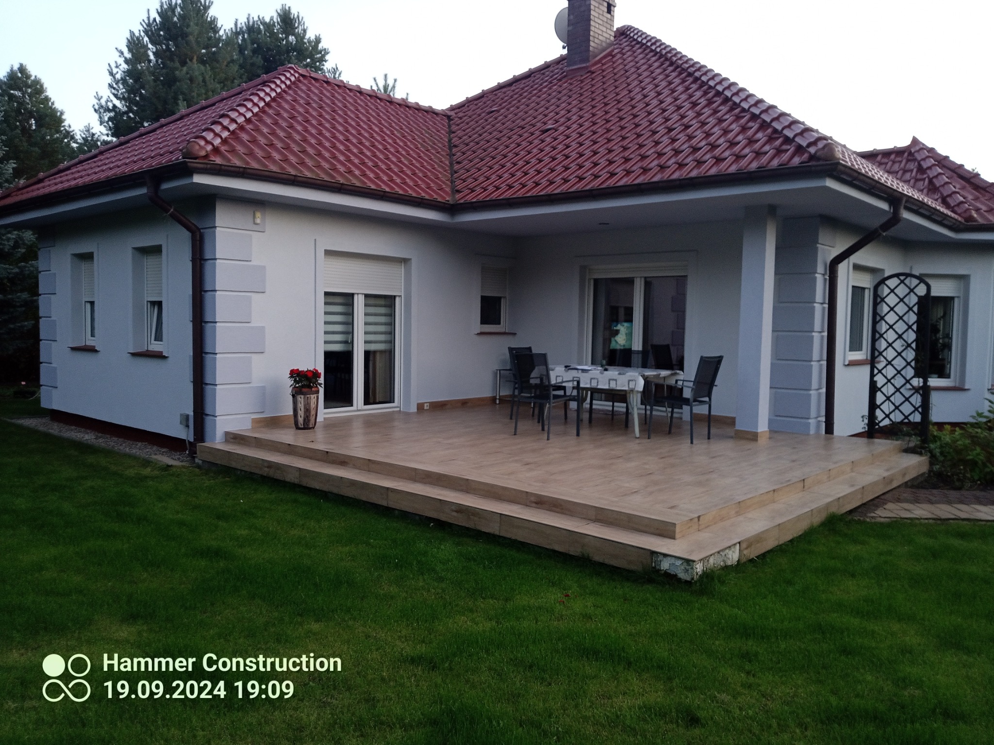 Dom z czerwonym dachem i jasną elewacją, widok z ogrodu. Taras z drewnopodobnych płytek, krzesła i stół na zewnątrz. Uwagę przykuwają detale wykończenia elewacji.