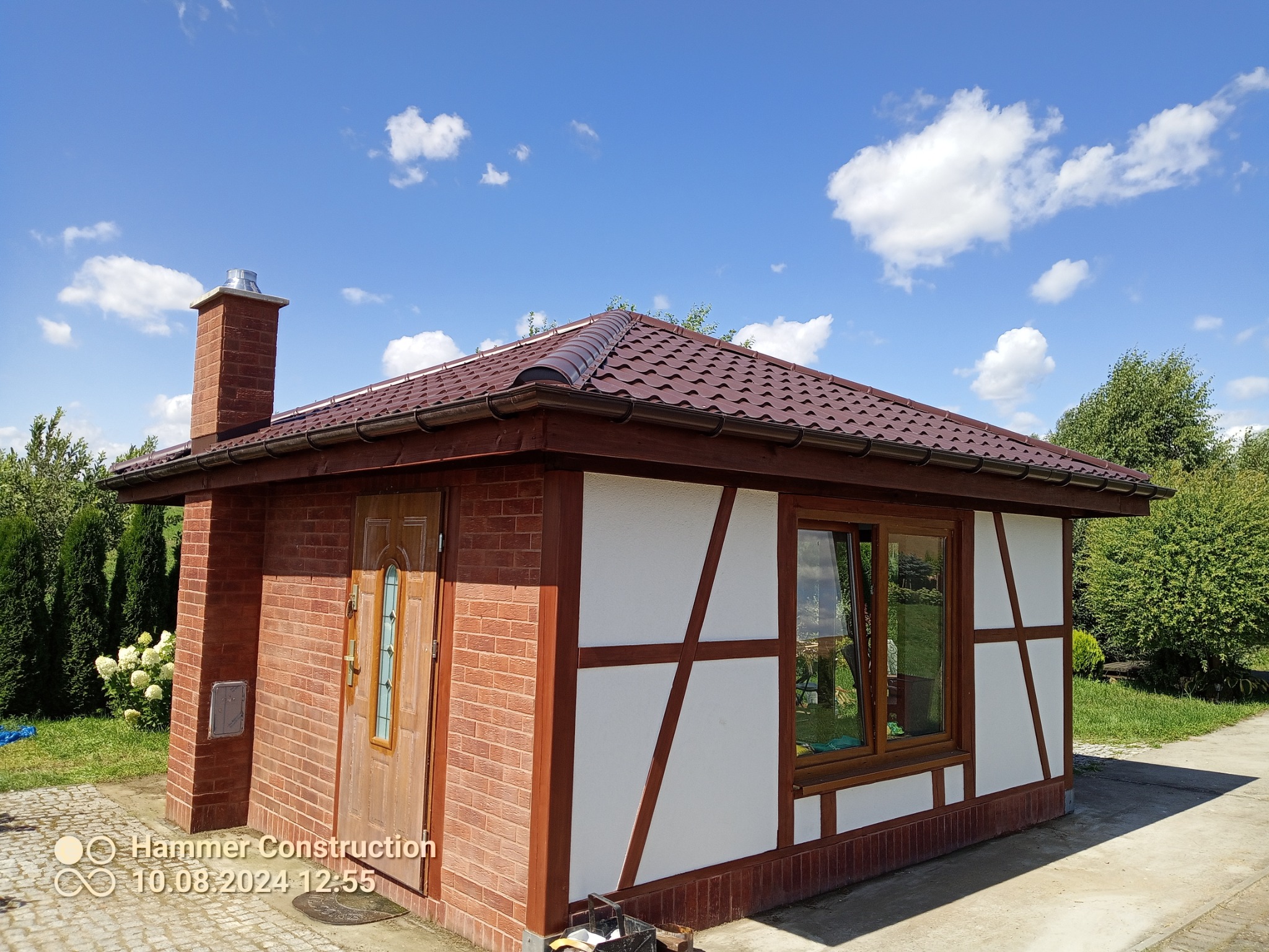 Wykończony budynek w stylu muru pruskiego z brązowym dachem, kominem i stolarką okienną na tle błękitnego nieba z chmurami, otoczony zielenią.