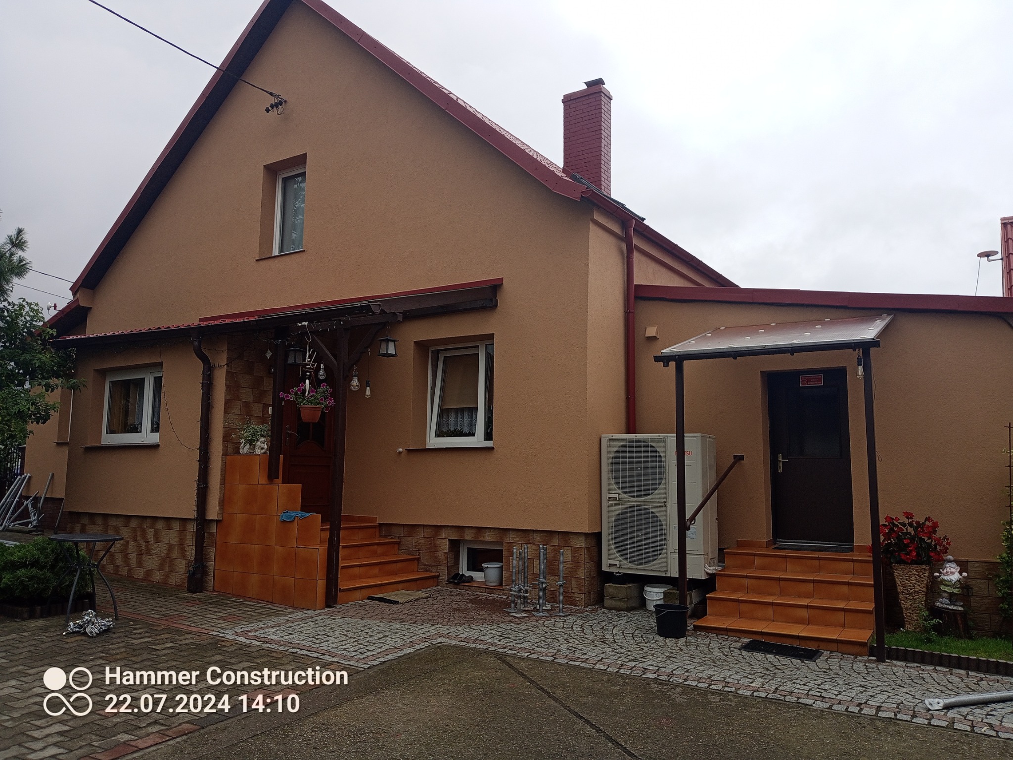 Dom jednorodzinny po remoncie elewacji w odcieniach beżu i brązu, z widocznym fragmentem dachu i komina. Na pierwszym planie podłoże z kostki brukowej.