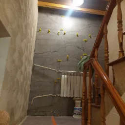 Ściana w trakcie układania płytek imitujących kamień, z widocznymi krzyżykami dystansowymi, grzejnik oraz drewniane schody w tle.