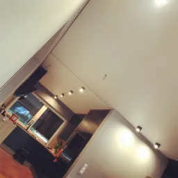 Nowoczesna kuchnia z drewnianą podłogą, szarymi ścianami i białym sufitem z zamontowanymi punktowymi lampami LED. Widoczne okno z roletami i zabudowa kuchenna.