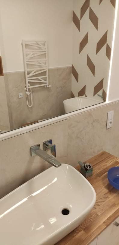 Nowoczesna łazienka z białą umywalką nablatową, chromowaną baterią ścienną i drewnianym blatem, widoczny fragment grzejnika drabinkowego i dekoracyjne płytki na ścianie.