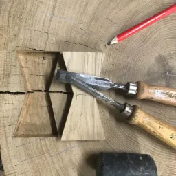 Drewniany blat z wyfrezowanym wzorem w kształcie klepsydry, obok dłuta, ołówek i gumowy młotek, praca w toku.