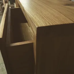 Otwarta szuflada z jasnego drewna z widoczną strukturą słojów, część szafki, ciepłe oświetlenie.