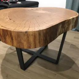 Stolik kawowy z blatem z naturalnego drewna, o nieregularnym kształcie i widocznych słojach, osadzony na metalowej, czarnej podstawie w kształcie litery X, na tle podłogi z płytek imitujących drewno.