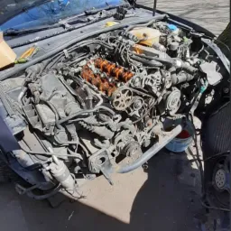 Otwarta maska samochodu z widocznym, zdemontowanym silnikiem; pomarańczowe wałki rozrządu, pasek rozrządu i inne elementy mechaniczne w warsztacie.