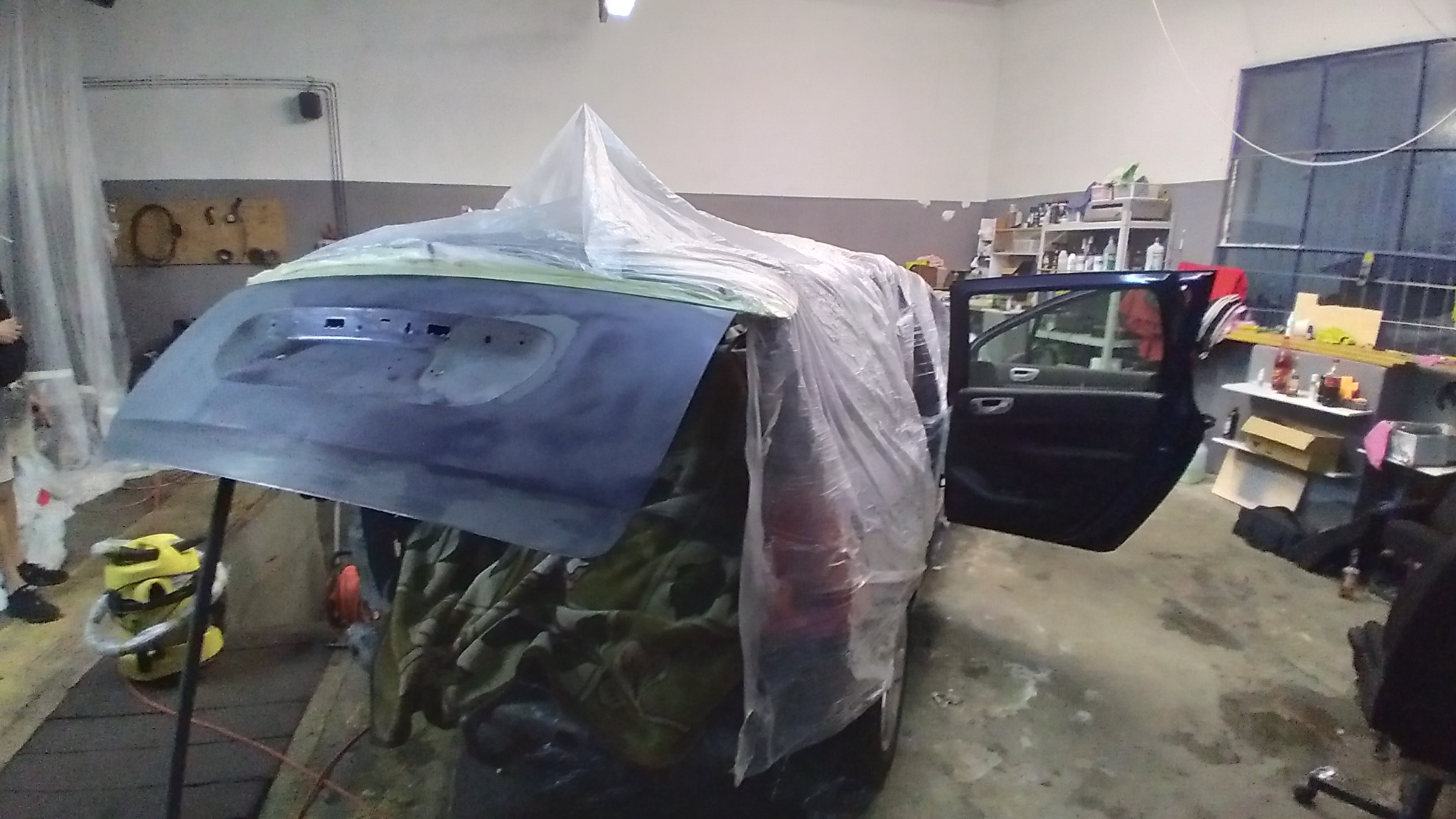 Samochód przykryty folią w warsztacie podczas przygotowania do lakierowania, widoczny podkład na klapie bagażnika, otwarte drzwi i wyposażenie warsztatu w tle.