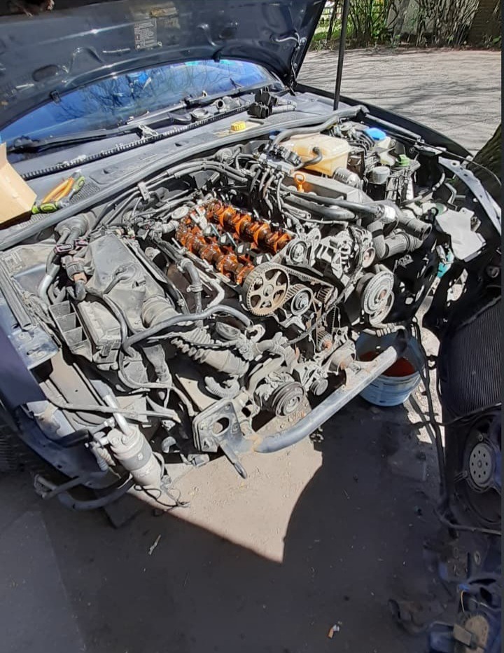 Otwarta maska samochodu z widocznym, zdemontowanym silnikiem; pomarańczowe wałki rozrządu, pasek rozrządu i inne elementy mechaniczne w warsztacie.