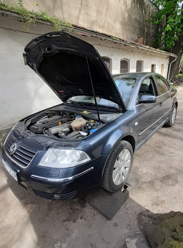 Granatowy Volkswagen Passat z otwartą maską, silnik widoczny, zaparkowany na betonowej płycie przed budynkiem z białego tynku.