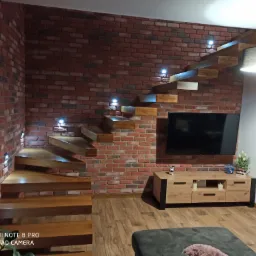 Nowoczesne schody drewniane, wspornikowe, zamontowane na ceglanej ścianie z punktowym oświetleniem, widok na salon z telewizorem.