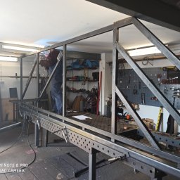 Metal Project - Pracownik w kombinezonie spawalniczym łączy metalowe elementy konstrukcji spawarką w warsztacie, widoczne iskry i narzędzia na ścianie.