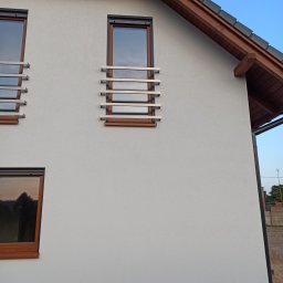Metal Project - Elewacja budynku z trzema oknami zabezpieczonymi poziomymi, metalowymi kratami o prostym, nowoczesnym designie, z drewnianymi ramami okiennymi i krokwiami dachu.