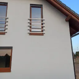 Elewacja budynku z trzema oknami zabezpieczonymi poziomymi, metalowymi kratami o prostym, nowoczesnym designie, z drewnianymi ramami okiennymi i krokwiami dachu.