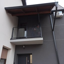 Metal Project - Nowoczesny balkon z balustradą szklaną i pionowymi metalowymi elementami, zadaszony drewnianą konstrukcją, na tle szarej elewacji domu.