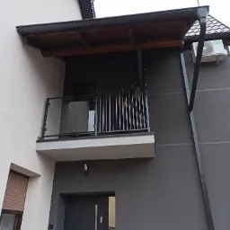 Nowoczesny balkon z balustradą szklaną i pionowymi metalowymi elementami, zadaszony drewnianą konstrukcją, na tle szarej elewacji domu.