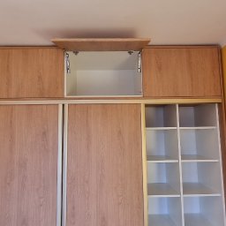 Tomasz Kuczynski - Wysoka szafa wnękowa z przesuwnymi drzwiami o jasnym, drewnianym wykończeniu, z otwartą szafką górną na zawiasach i widocznymi półkami po bokach.