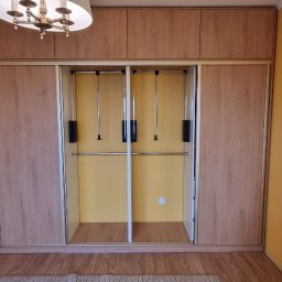 Tomasz Kuczynski - Szafa wnękowa z przesuwnymi drzwiami i systemem pantografów w Ostrowi Mazowieckiej, z otwartą częścią regałową po lewej stronie, na tle żółtej ściany.